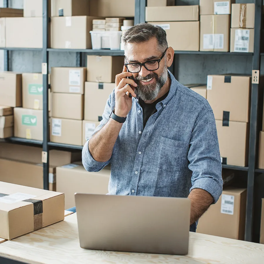 A man speaking on the phone while looking at his laptop in a warehouse.